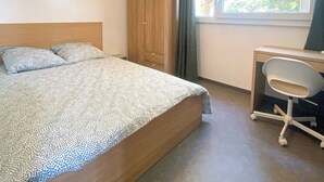 Ein Schlafzimmer mit einem Bett, einem Kleiderschrank, einem Schreibtisch mit Stuhl und einem Fenster mit Blick auf Bäume.