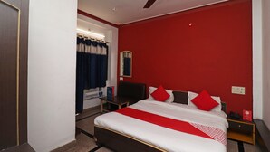 Quarto de hotel com cama, mesa de cabeceira, escrivaninha, ventoinha no teto e janela com cortinas.