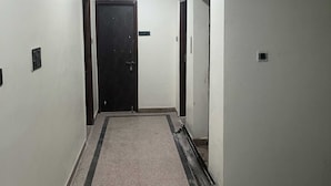 Um corredor estreito com uma porta escura no final, paredes brancas e chão de azulejos.