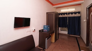 Quarto de hotel com um sofá marrom, uma pequena mesa de trabalho com uma televisão e uma caixa, um guarda-roupa e uma janela com cortinas azuis.
