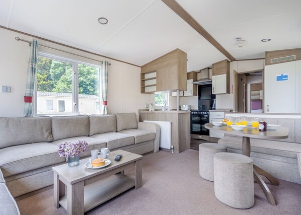 A modern, open-plan living space with a sofa, coffee table, and ottoman, a kitchen area with appliances, and a dining table set with breakfast. - Gold Caravan Accessible. With parking and decking. (Ryde, Nr Bembridge)