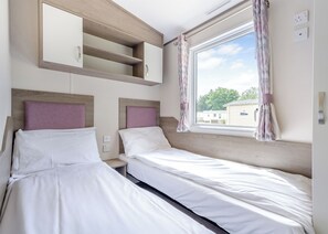 A bedroom with two beds, built-in shelves, a window with curtains, and a view of the outside. - Gold Caravan Accessible. With parking and decking. (Ryde, Nr Bembridge)