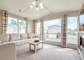 A living room with a sofa, a coffee table, and a television. There are large windows with curtains, and a ceiling fan is visible. - Gold 2 Bedroom Caravan, Sleeps 6. With decking. (Walton-on-the-Naze)