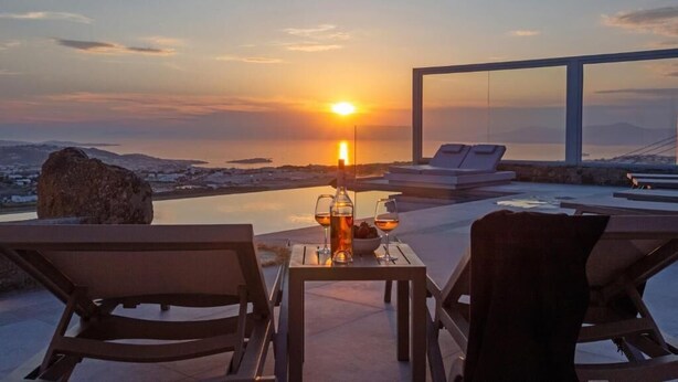 Een zonsondergang vanaf een terras met loungebedden, een tafel met een fles en glazen, en een heldere lucht.