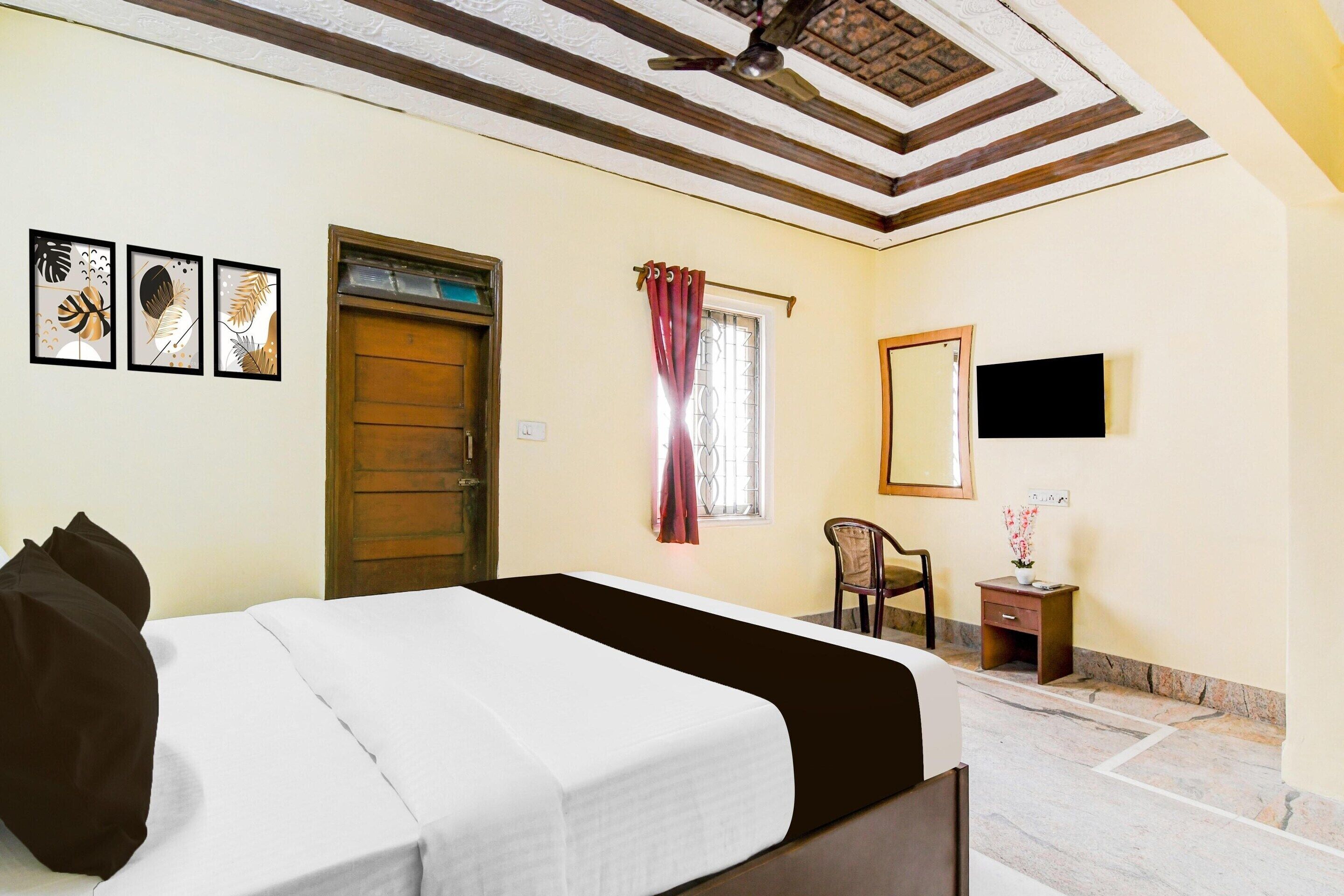 Quarto com cama, porta de madeira, janela com cortinas, uma cadeira, mesa pequena com um vaso e ventilador de teto.