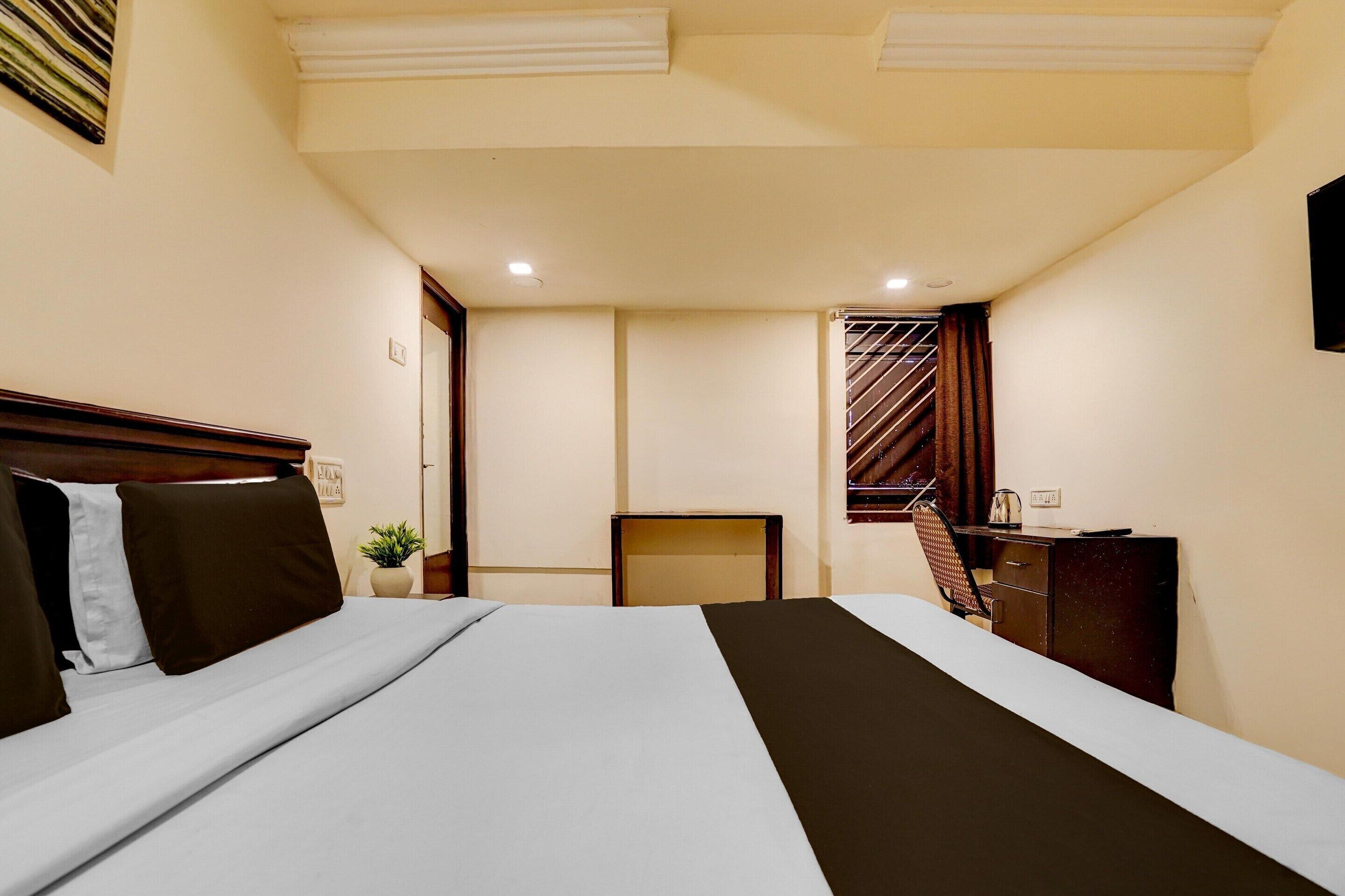 Una habitación de hotel con cama, escritorio, silla, planta en maceta y ventana con cortinas.