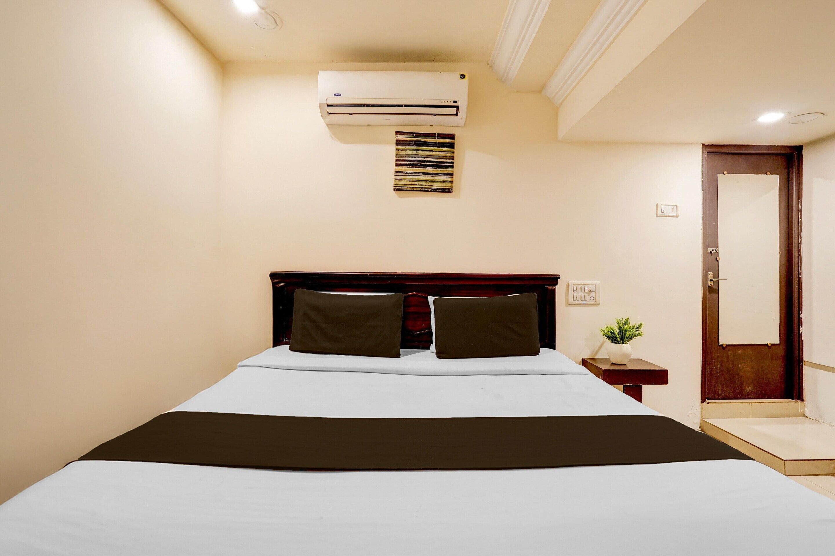 A hotel room with a bed, an air conditioner, a small bedside table with a plant, and a door.