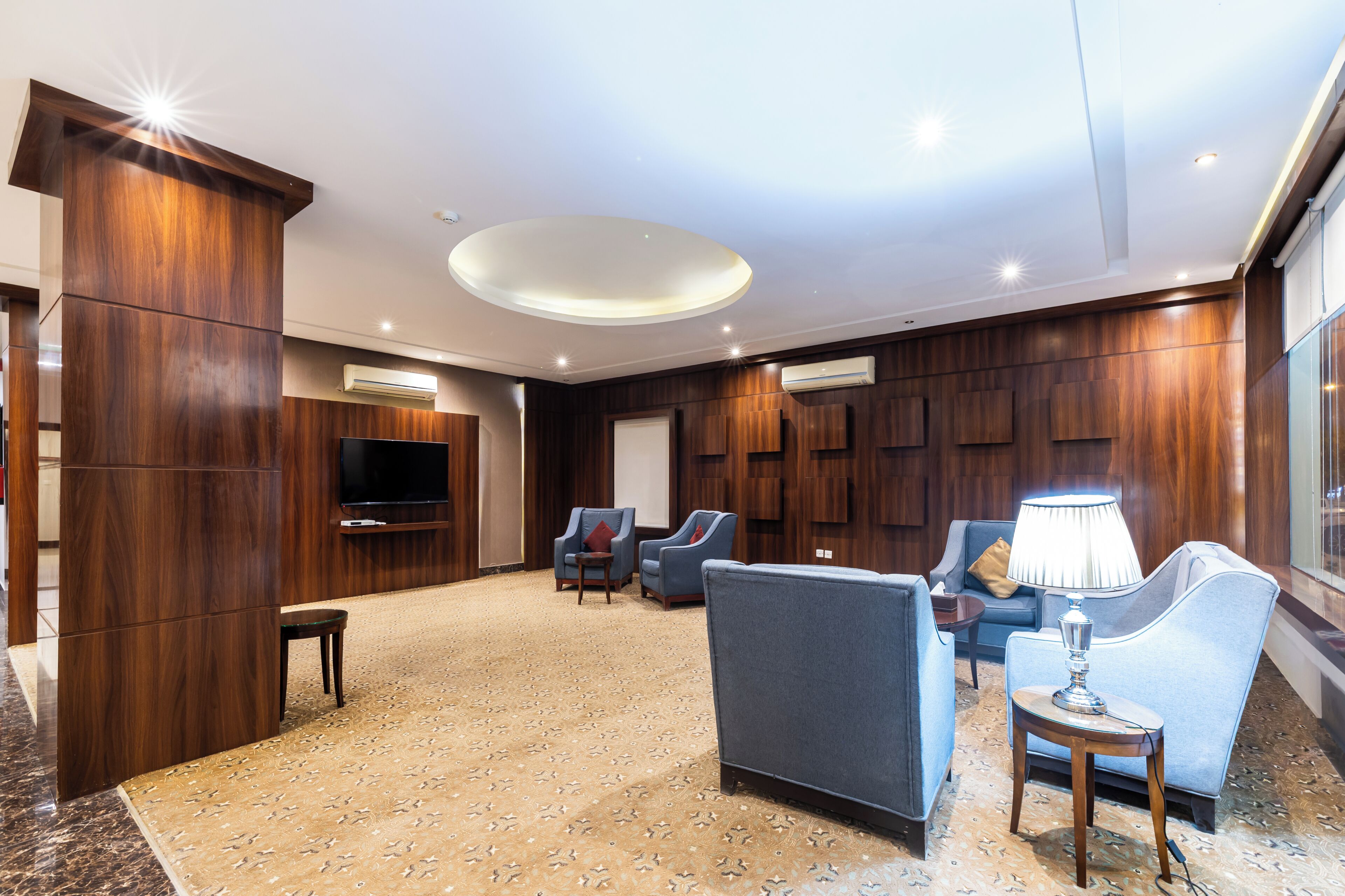 A spacious lobby with a high ceiling, a flat-screen TV mounted on a wooden panel, multiple seating areas with armchairs and sofas, and a patterned carpet.