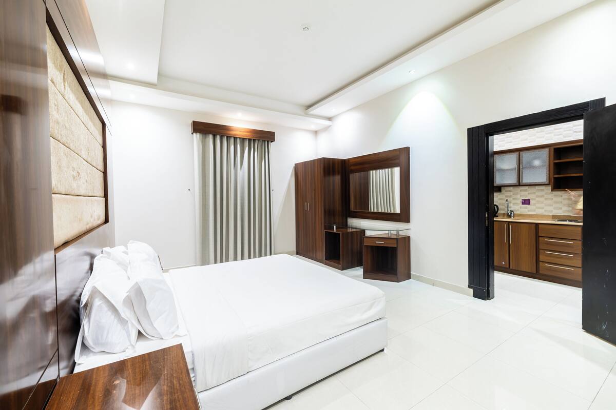Quarto de hotel moderno com cama, escrivaninha, espelho e cozinha compacta.