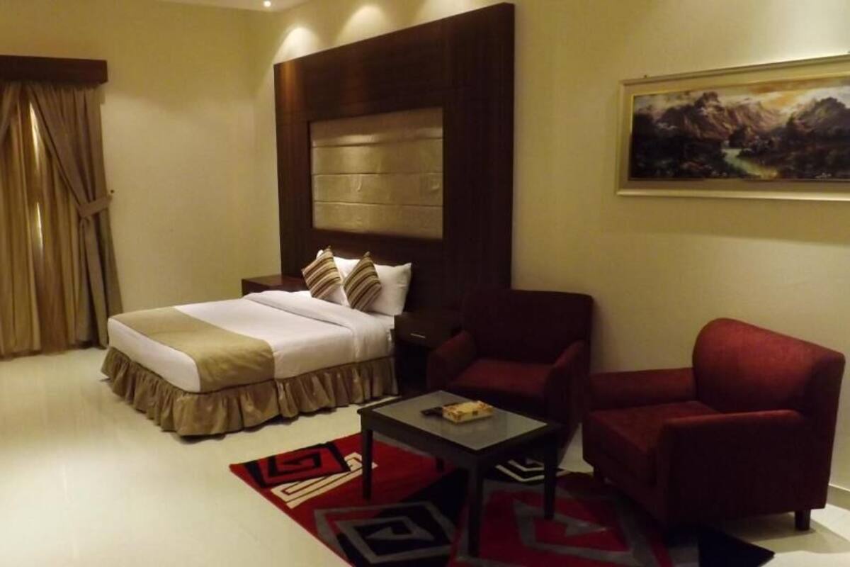 Quarto de hotel com uma cama, uma poltrona vermelha, uma mesa pequena, um quadro na parede e uma janela com cortinas.