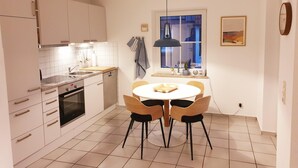 Et moderne køkken med hvide skabe, en indbygget ovn, et spisebord med fire stole og et vindue med udsigt til en bygning.