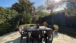 Een terras buiten met een tafel en stoelen, omgeven door groen en een gebouw op de achtergrond.