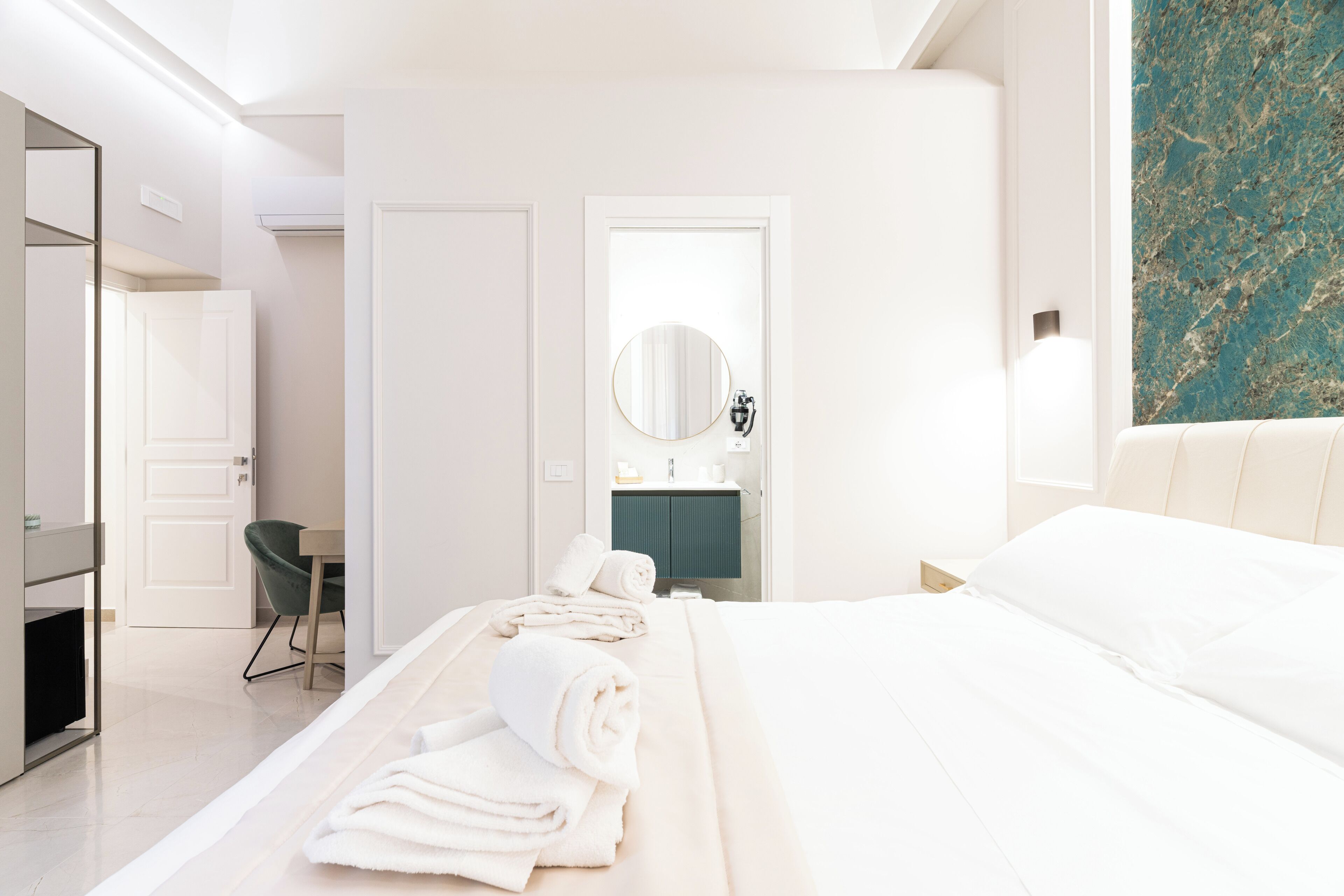 A modern hotel room with a bed, a desk, a chair, a mirror, and a bathroom visible through an open door.