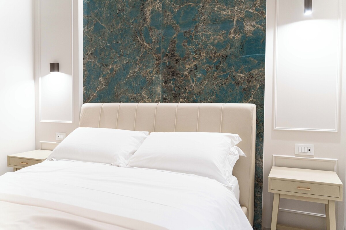 Un lit bien fait, avec du linge de lit blanc, un mur décoré d’un motif marbré derrière, et deux tables de chevet avec des lampes.