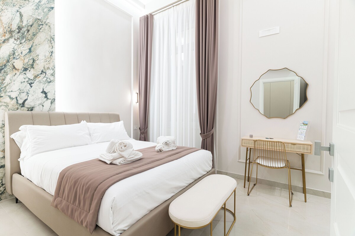 Une chambre d’hôtel comprenant un lit, une table de chevet, une chaise, un miroir et un mur orné d’un motif rappelant le marbre.