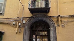 A yellow building with a metal gate, a balcony with plants, and a sign on the door. - Nimbus Rooms (Naples)