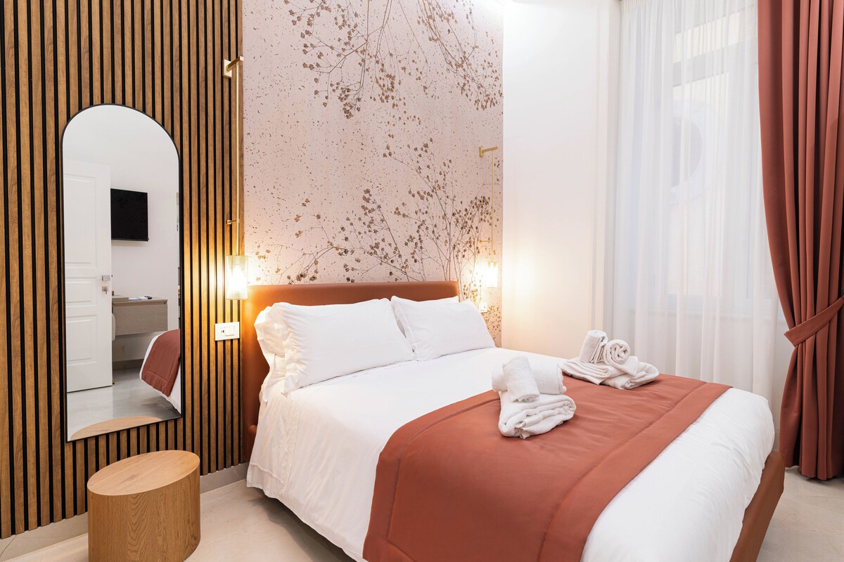 Une chambre d’hôtel moderne avec un grand lit, une petite table d’appoint ronde en bois, un miroir et un téléviseur fixé au mur.