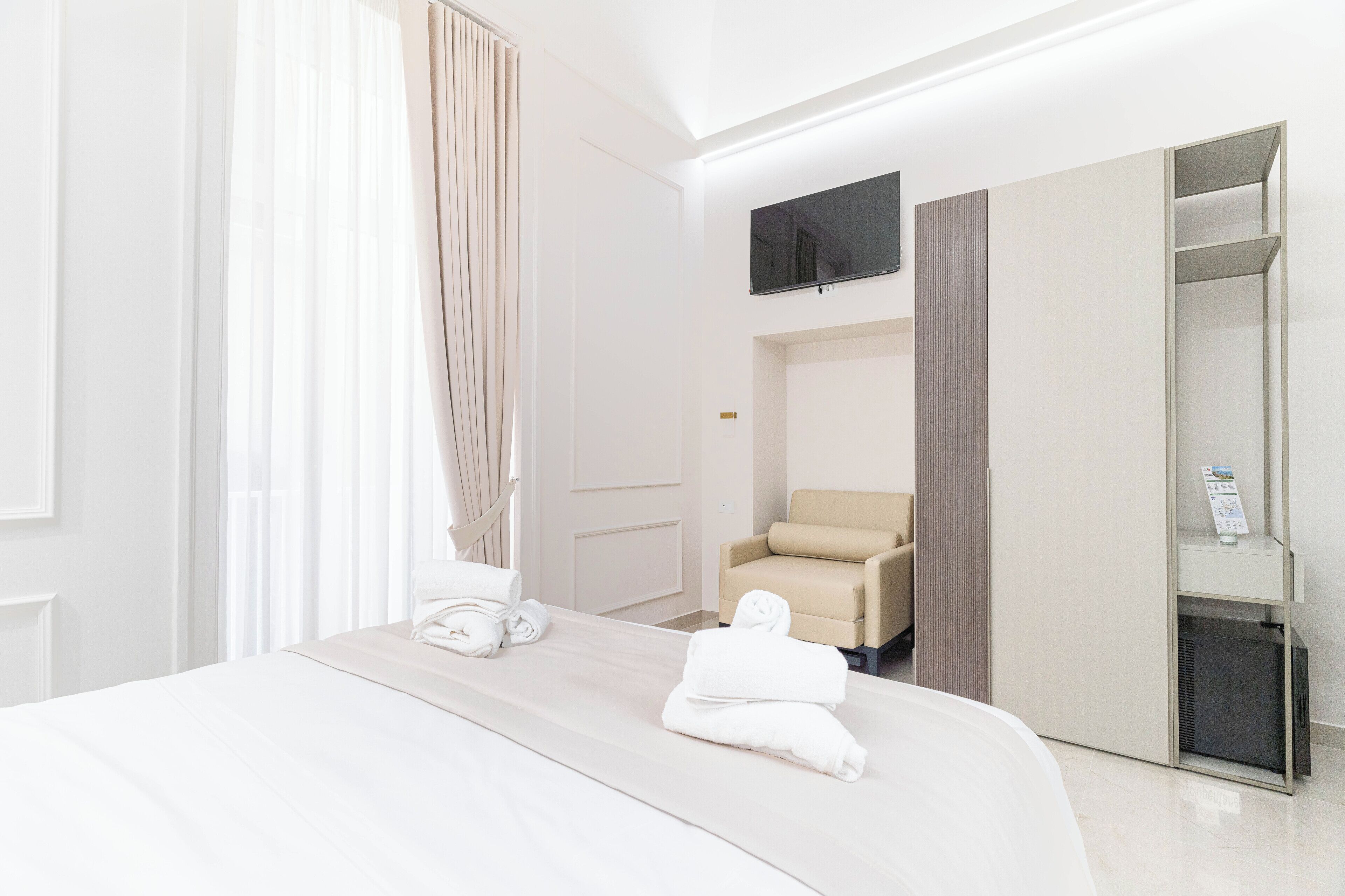 A hotel room with a bed, a chair, a TV, and a minibar.