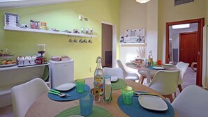 Zona pranzo con tavolo apparecchiato per quattro, circondato da sedie bianche. Sopra il tavolo, scaffali con diversi oggetti, e si intravede un frigorifero sullo sfondo.