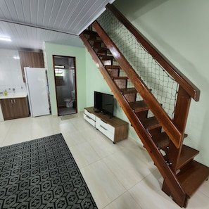 Ein Wohnzimmer mit einer Holztreppe, einem Fernseher auf einem Holzboden, einer Küchenzeile und einem gemusterten Teppich.