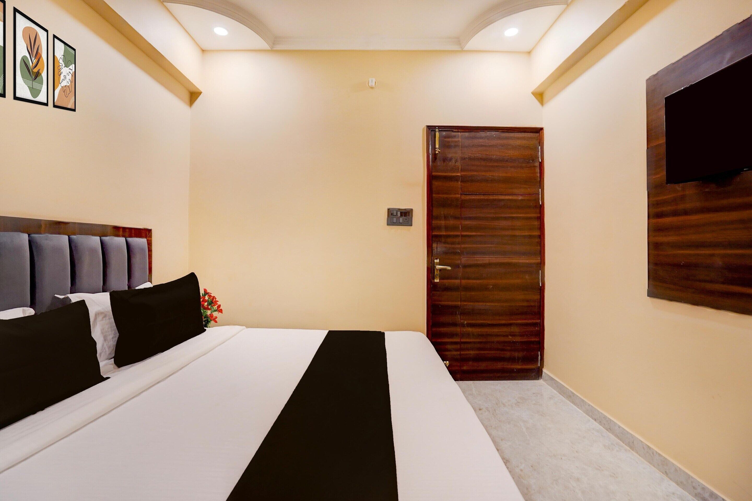 A hotel room with a bed, a wooden door, a flat-screen TV mounted on the wall, and a ceiling light fixture.