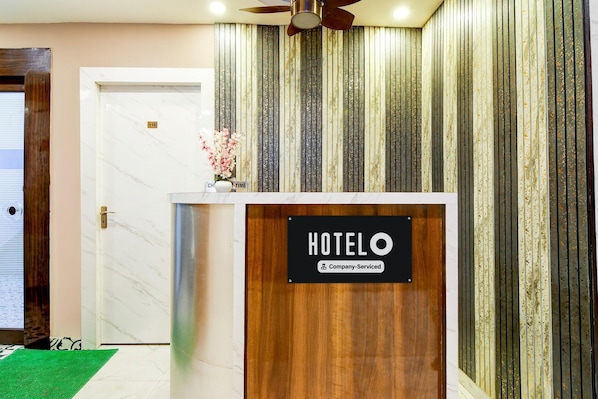 Un comptoir d’accueil d’hôtel avec une pancarte indiquant qu’il est géré par l’entreprise, une porte sur la gauche et un tapis vert sur le sol.