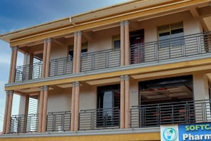Un immeuble à plusieurs étages doté d’un balcon au rez-de-chaussée, de colonnes et d’une enseigne indiquant « PHARMACIE SOFTC ».