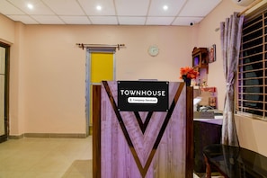 Zona de recepción con un cartel que dice 'TOWNHOUSE', un reloj, una ventana con cortinas y un escritorio con un jarrón de flores.