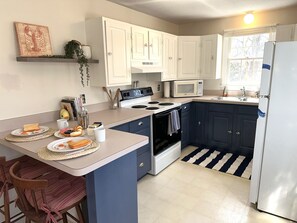 A kitchen with white cabinets, a microwave, stove, and a refrigerator. There are two bar stools and a small island with plates of food.
