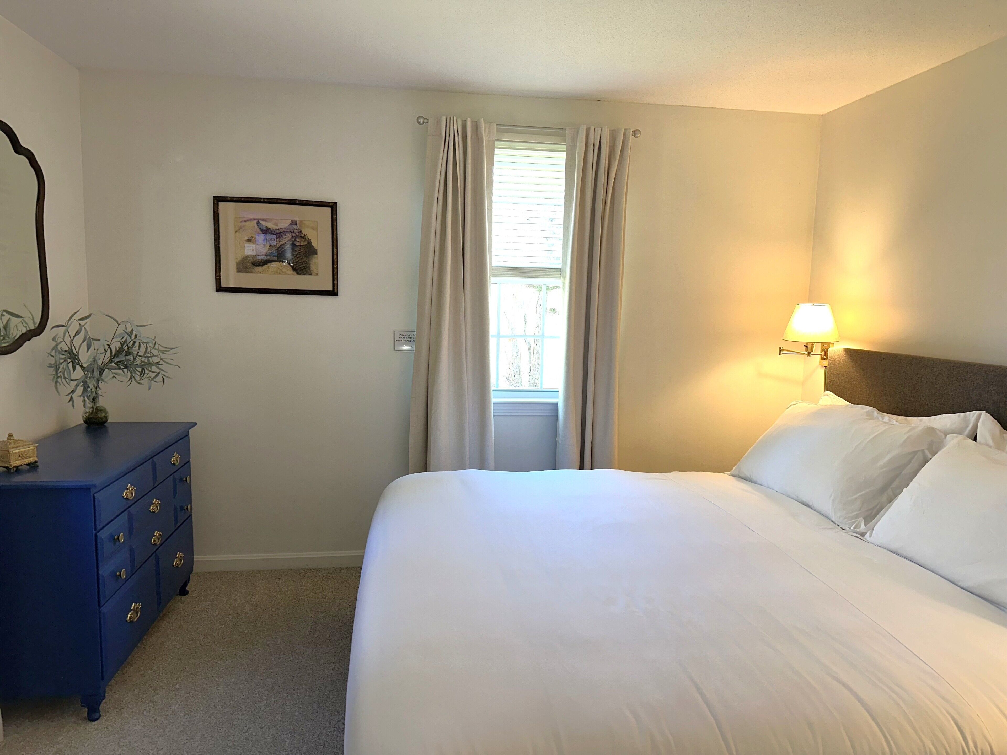 A bedroom with a bed, a dresser, a lamp, a window with curtains, and a framed picture on the wall.
