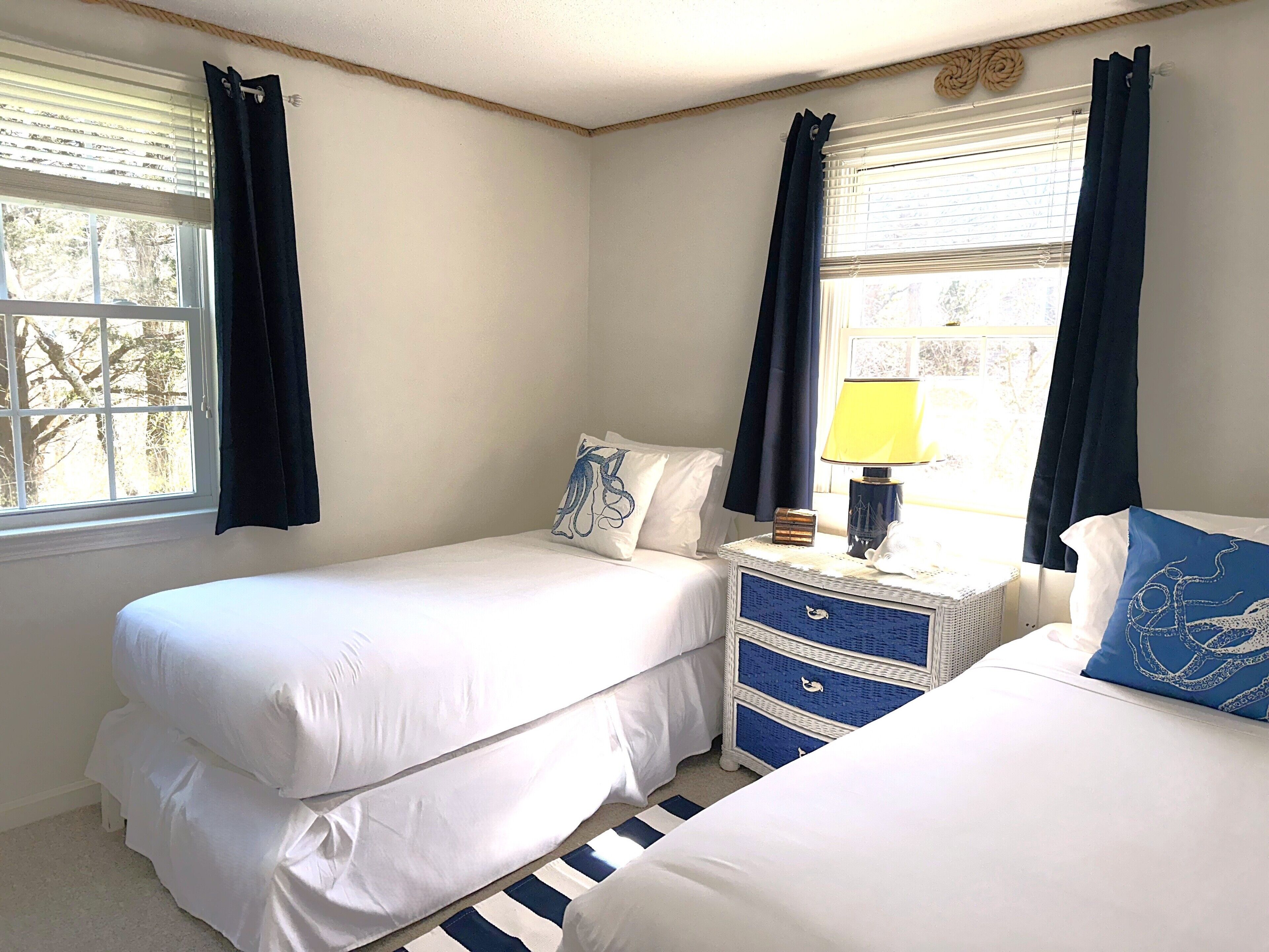 A bedroom with two beds, a nightstand with drawers, a lamp, and a window with curtains.