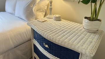A neatly made bed with white linens, a bedside lamp, a small white container, and a potted plant on a wicker nightstand.