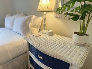A neatly made bed with white linens, a bedside lamp, a small white container, and a potted plant on a wicker nightstand.