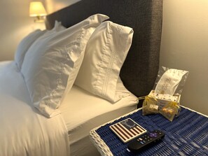 Uma cama bem arrumada com lençóis brancos, um controle remoto, uma pequena sacola de presente e um objeto decorativo sobre um descanso tecido azul.