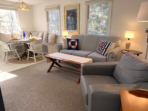 Uma sala de estar com um sofá cinzento, uma mesa de centro de madeira e um espaço de jantar com cadeiras e uma mesa.