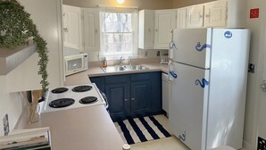 Une cuisine de taille réduite avec des meubles blancs, une cuisinière, un réfrigérateur et une fenêtre équipée de stores.