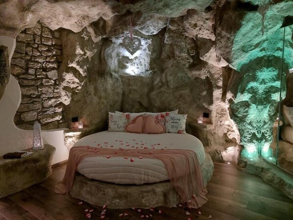 Une chambre avec un mur en pierre, un lit parsemé de pétales de rose et un projecteur diffusant une lumière verte sur le mur.