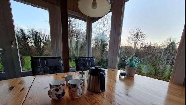 Una mesa de madera con tazas de café, un termo y una planta en maceta en una habitación con ventanas grandes y vistas a árboles.