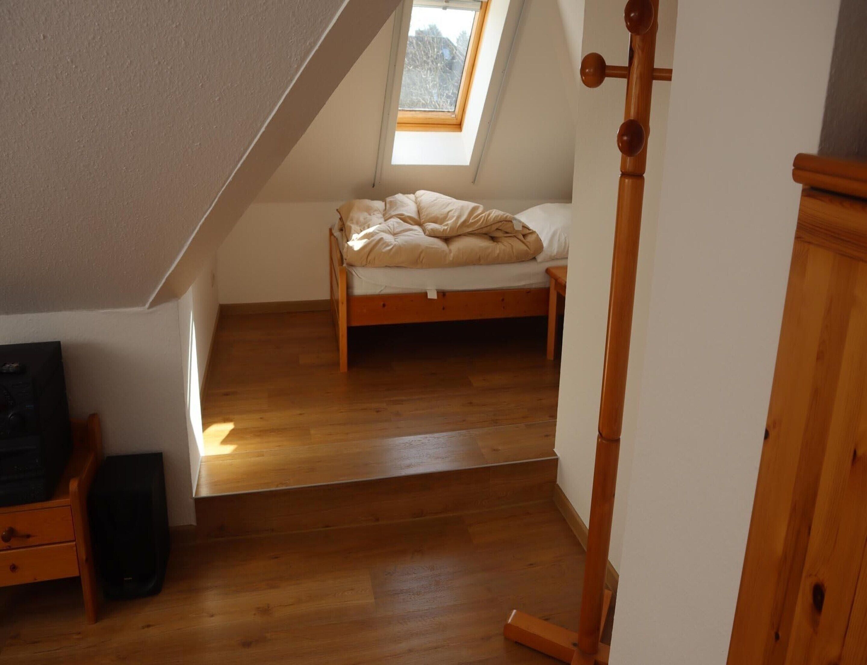 A room with a sloped ceiling, a bed with a wooden frame, a small table, and a speaker.