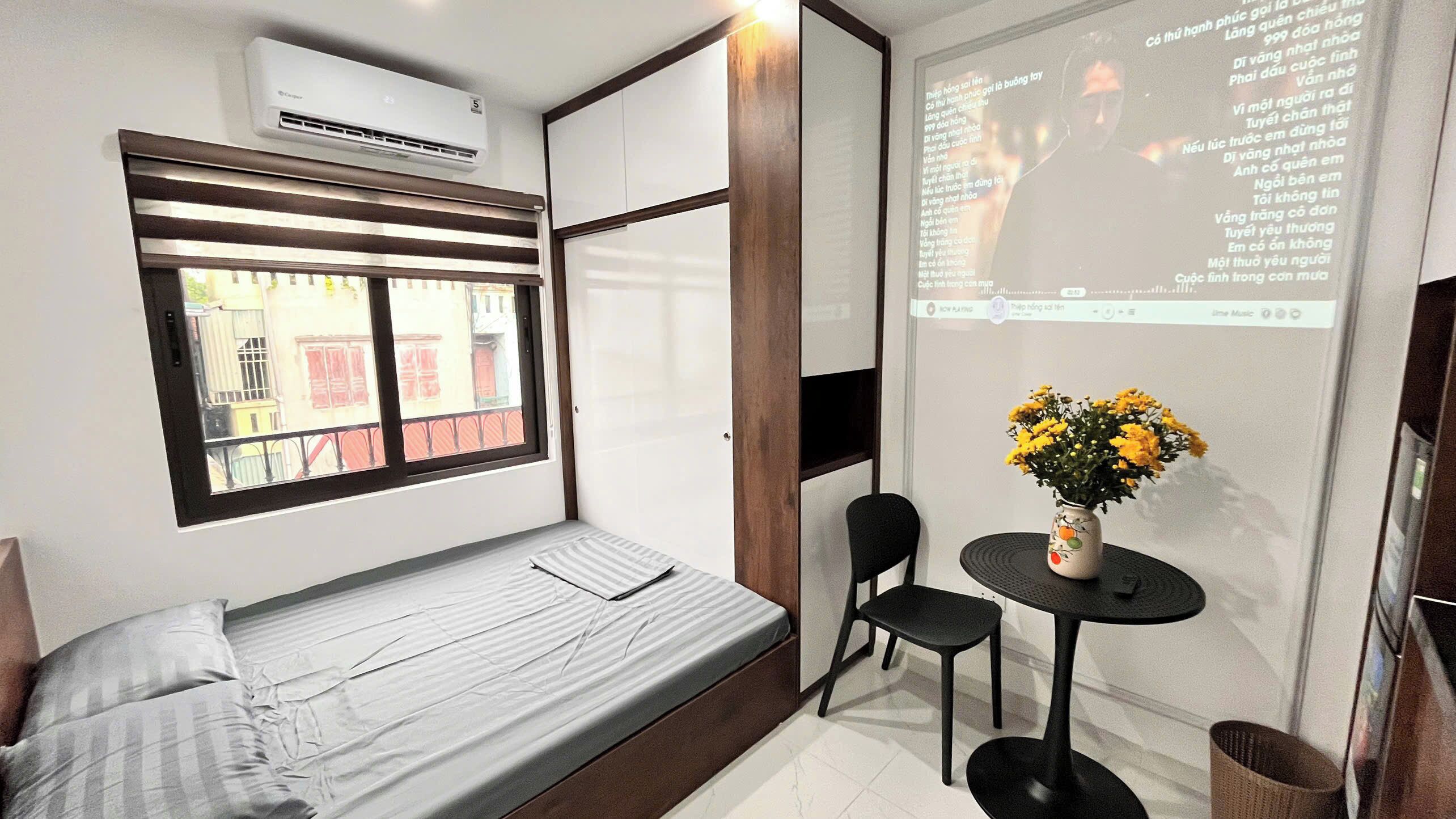 A small room with a bed, a window, a table with a vase of flowers, and a projection screen displaying text.