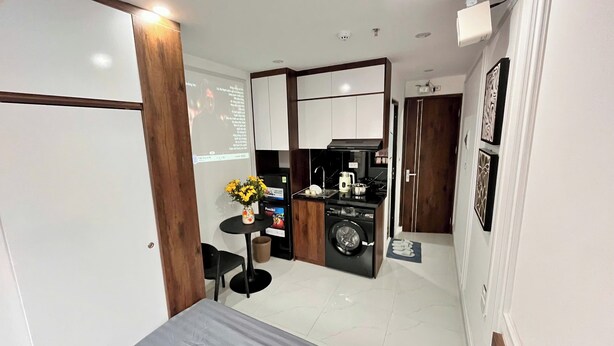 A compact hotel room with a kitchenette, a washing machine, a small table with a vase, and a bed. - Era Hong Mai Apartment (Hanoi)