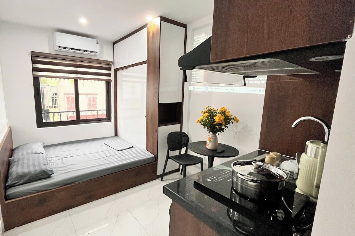 A compact room with a bed, a small table with a vase of flowers, a kitchen area, and a window with a view of buildings.