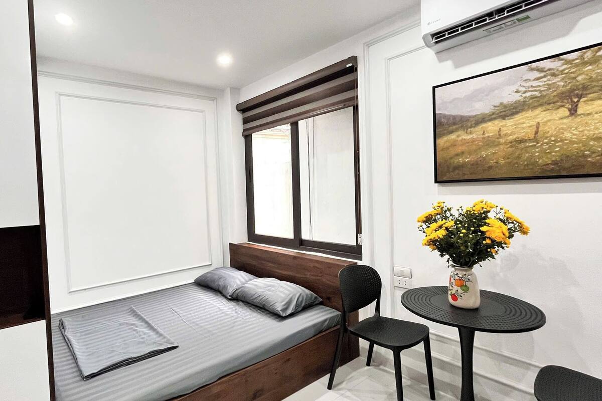 A compact room with a bed, a small table with chairs, a window with blinds, and a painting on the wall.