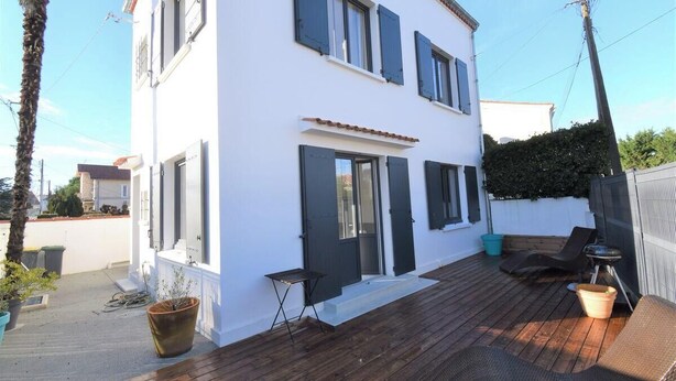 Un edificio blanco con contraventanas oscuras, una terraza de madera y mobiliario exterior.