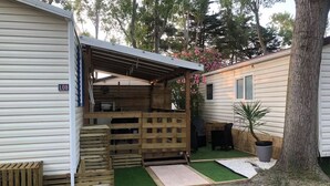 A mobile home with a covered outdoor area, a patio with a table and chairs, and a small garden with a tree and a potted plant. - L'Eden - Mobile home 2 bedrooms 6 people - Swimming pool Air conditioning - Campsite 5⭐️ (Le Grau-du-Roi)