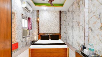A hotel room with a wooden bed, a ceiling fan, an air conditioning unit, a nightstand with a lamp, and a small table with a kettle and bottles.