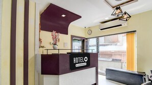 Een receptiebalie van een hotel met een bord waarop 'HOTELO Company-Serviced' staat, een klok, een vaas met bloemen en een raam met uitzicht.