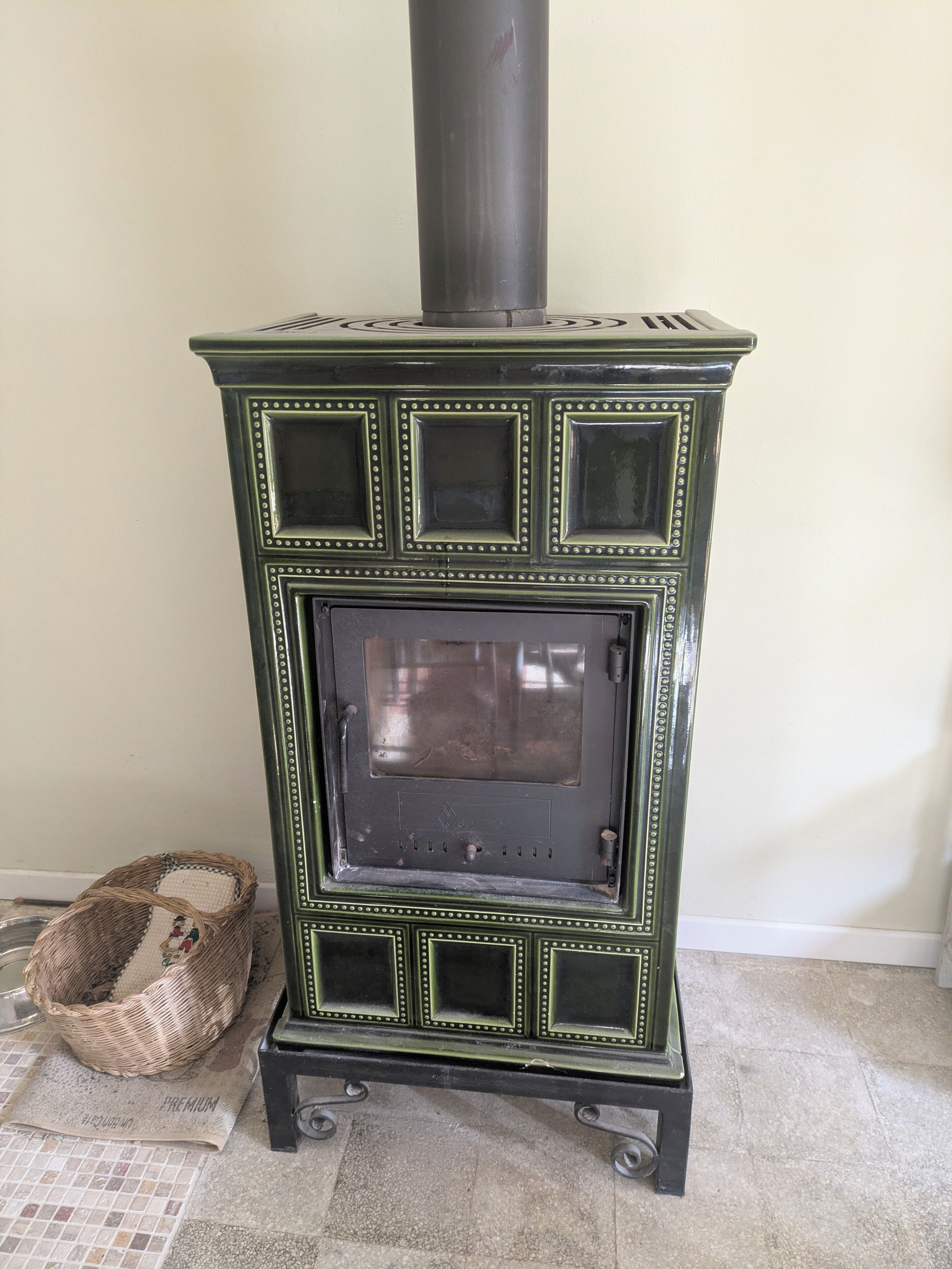 Estufa de madera estilo vintage verde con puerta de cristal, sobre un piso de baldosas, junto a una cesta tejida.