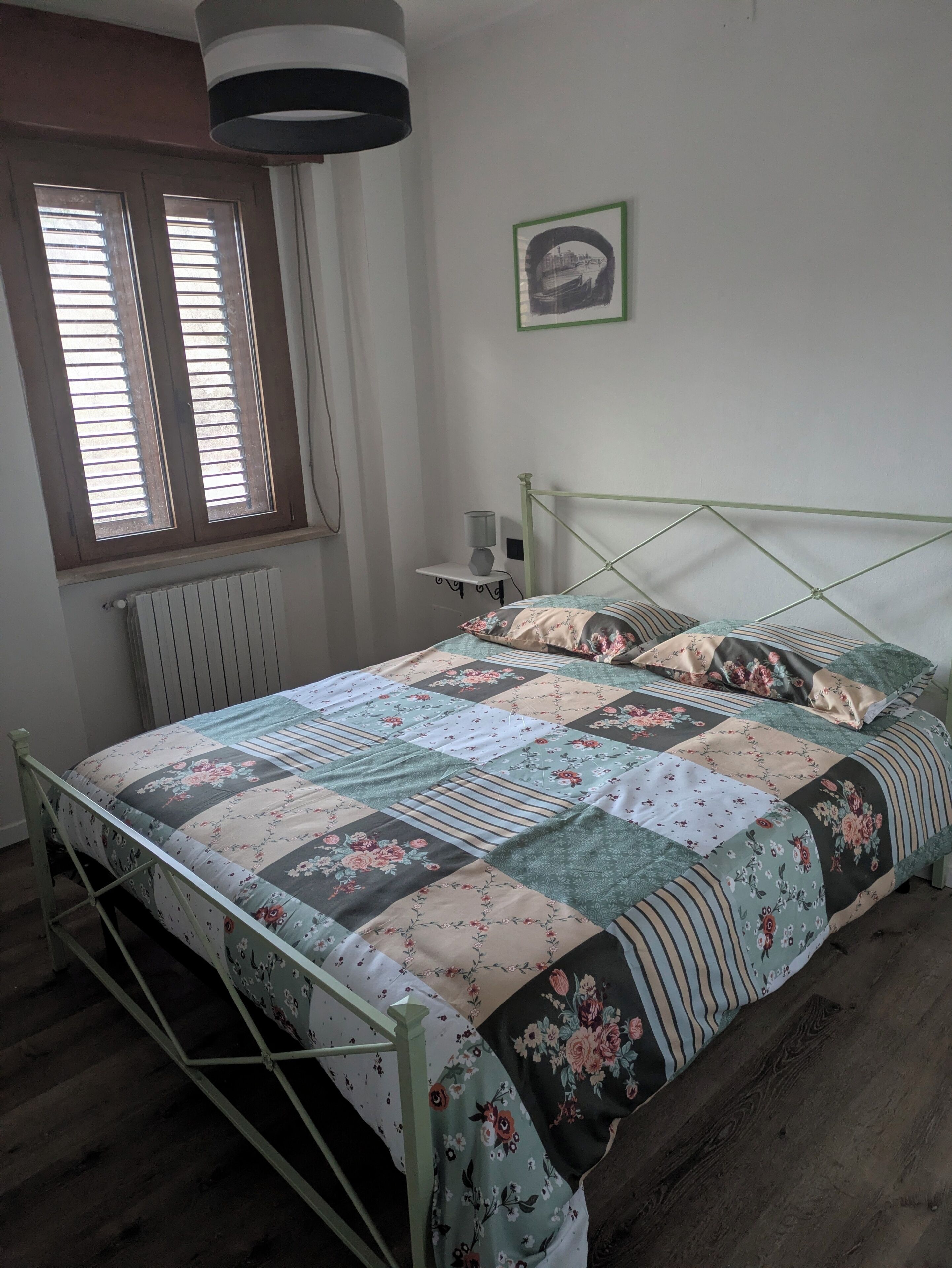 Quarto com estrutura de cama em metal, colcha acolchoada, radiador, janela com persianas e quadro fixado na parede.