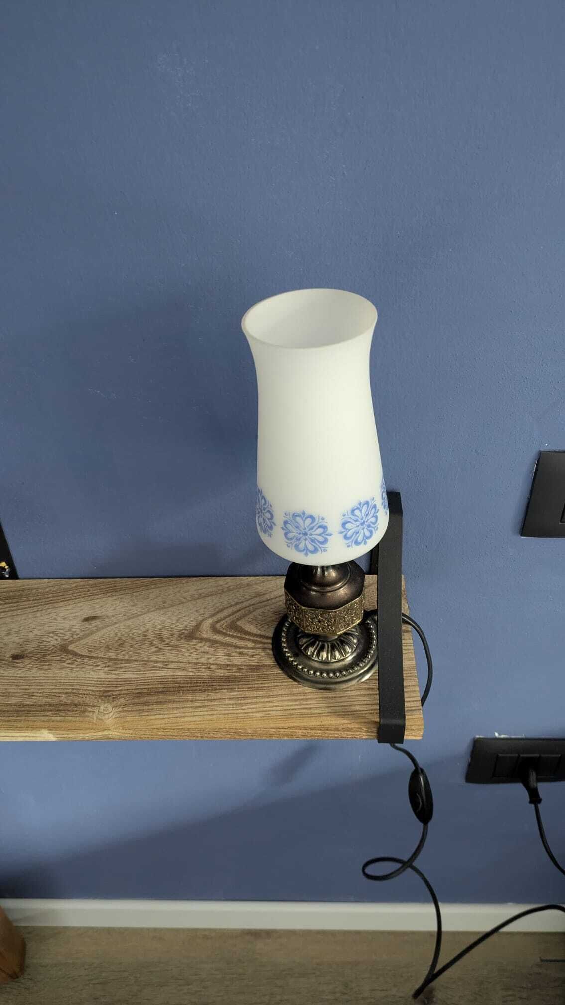 Una lampada bianca con motivi floreali blu su uno scaffale in legno, con uno sfondo azzurro.